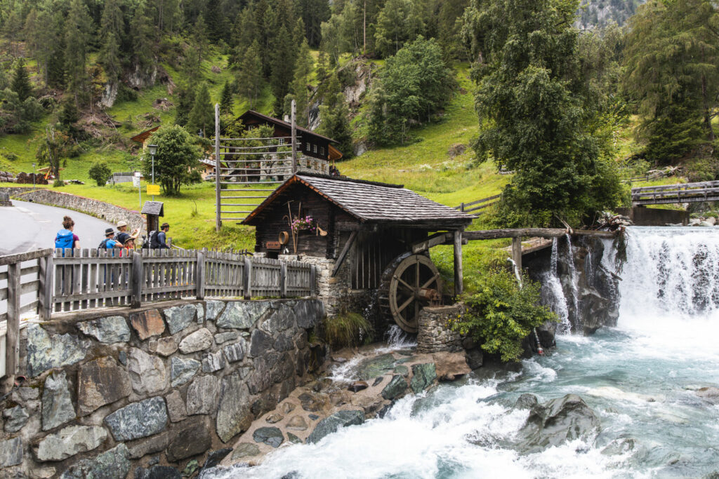 Eine kleine Mühle aus dunklem Holz neben der Isel. 4 Wandernde stehen daneben und betrachten sie. Im Hintergrund sieht man einen Bauernhof und Wald. Daneben verläuft eine Straße.
