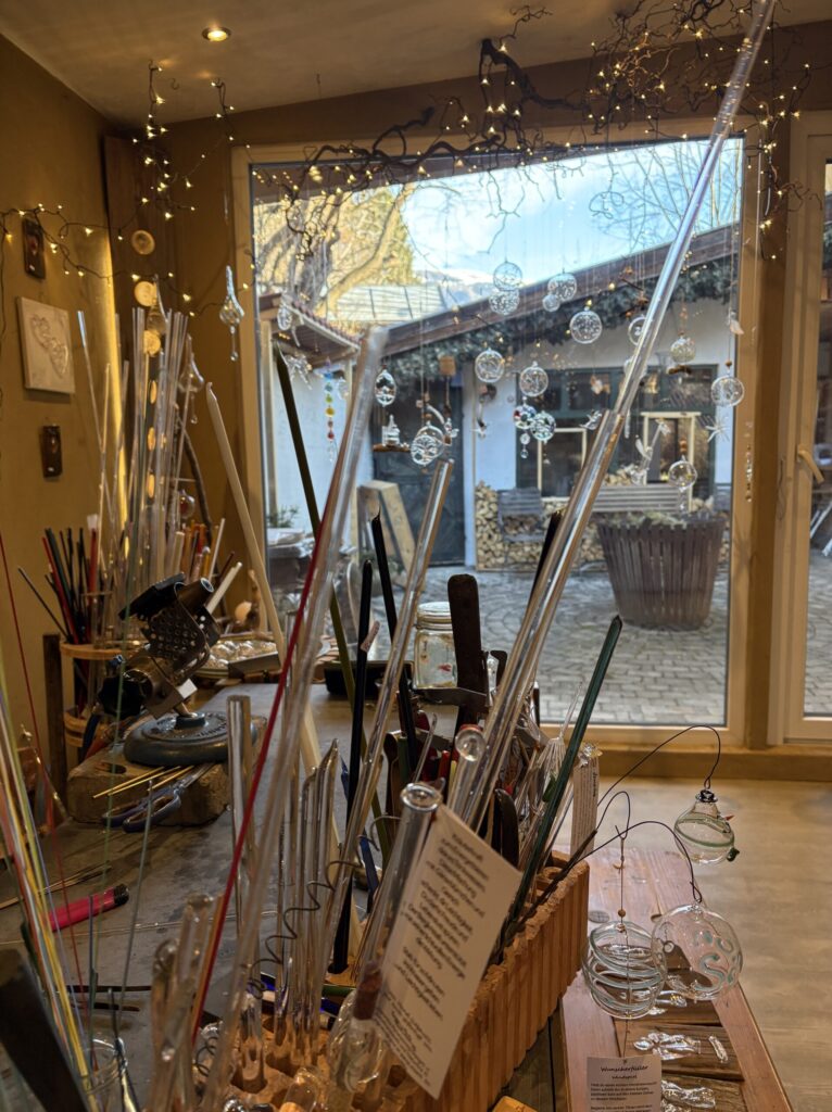 Bunte Glasröhren und Werkzeuge in der Glasgrün-Werkstatt. Im Hintergrund sind handgefertigte Glaskugeln und ein Innenhof zu sehen.