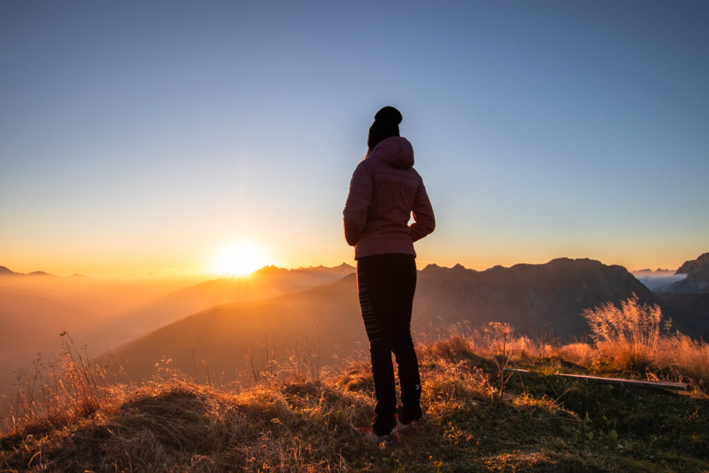 Eine Person steht alleine am Berg "Öfenspitze" und betrachtet den Sonnenaufgang und die in goldenes Licht getauchte Landschaft.