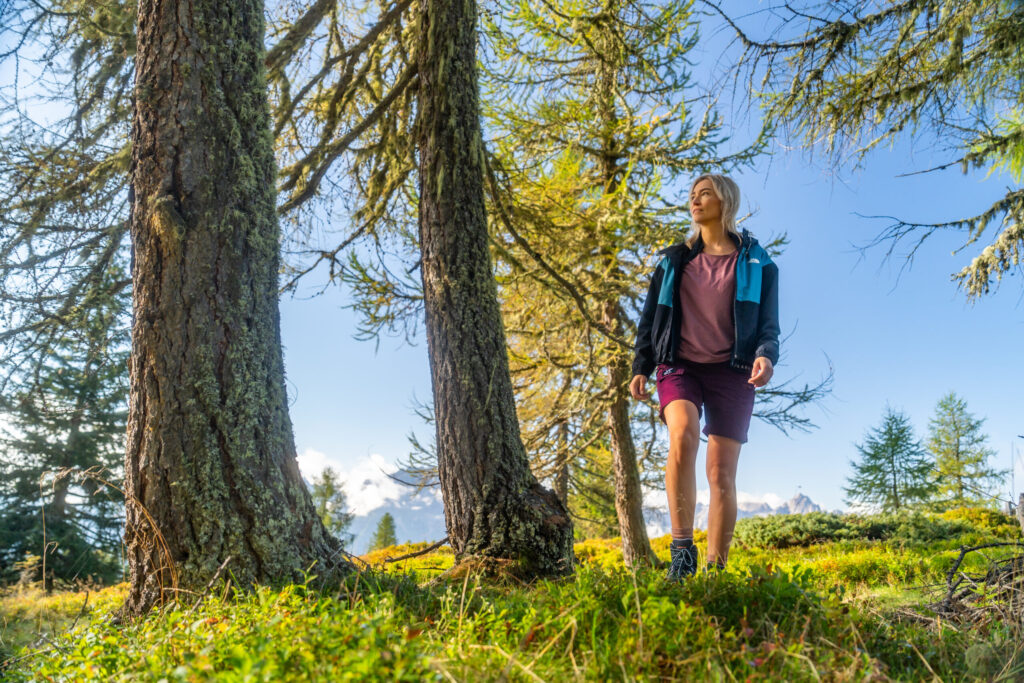 Eine blonde Frau mit sommerlicher Sportkleidung wandert hoch oben und allein am Berg durch einen Wald in Osttirol.