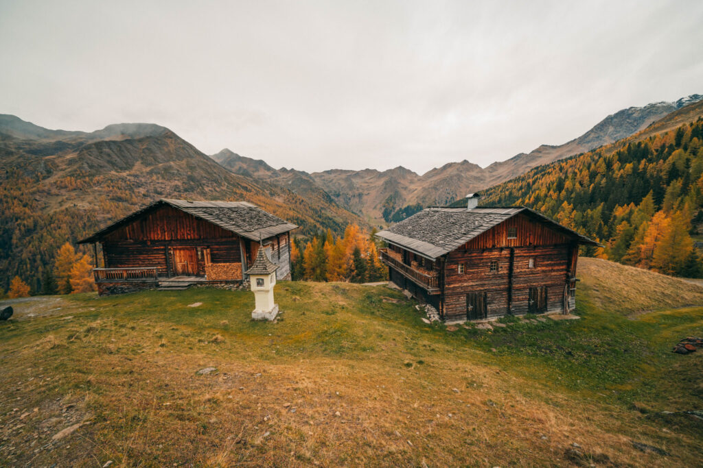 Zwei hölzerne Almhütten auf der herbstlichen Karmelisenalm, im Hintergrund stehen goldene Lärchen, grüne Fichten und das Bergpanorama des Villgratentals in Osttirol. Der perfekte Ort zum Bergbaden.