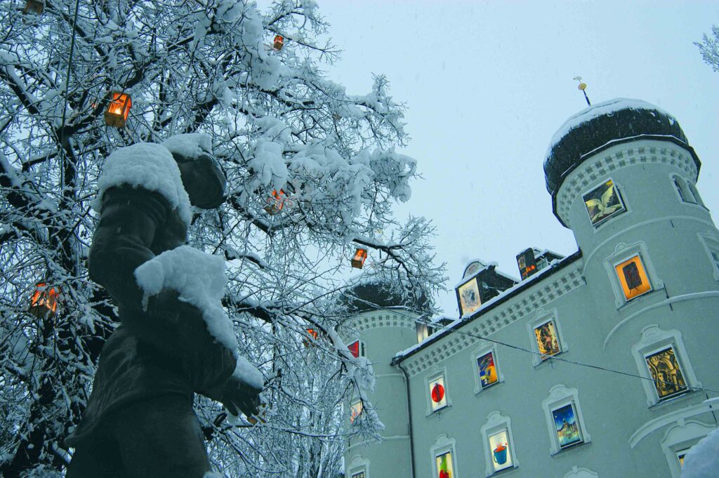Im Vordergrund ist die Statue auf dem Brunnen des Hauptplatz in Lienz, daneben befindet sich ein Baum mit Laternen. Im Hintergrund sieht man den Adventkalender der Liebburg. Es schneit und alles ist bereits mit Schnee bedeckt.