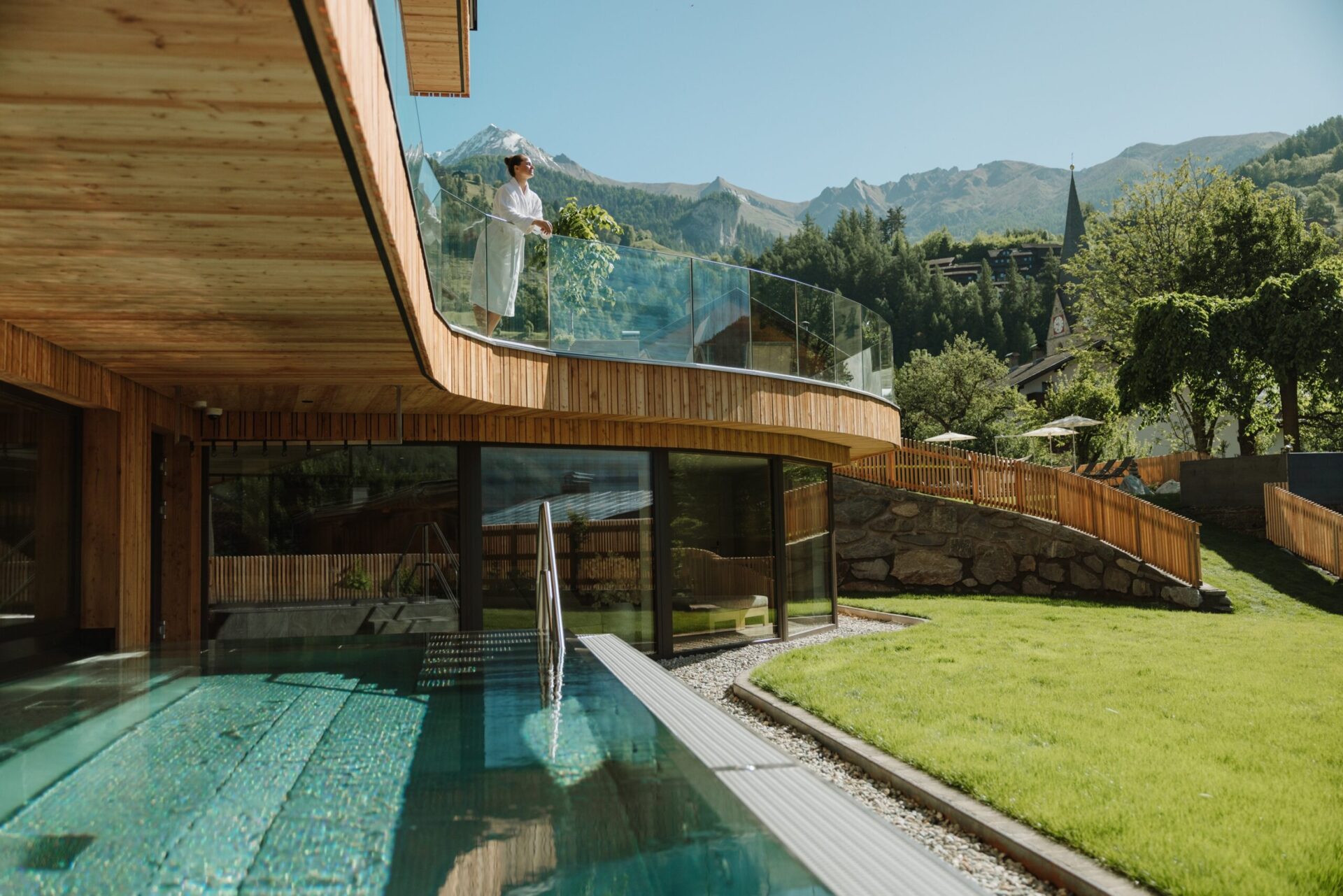 Ansicht vom Außenbereich des Naturhotel Outside. Eine Frau steht im weißen Bademantel auf einem Balkon und genießt die Sonne. Darunter befindet sich der Außenpool und im Hintergrund befinden sich Berge.
